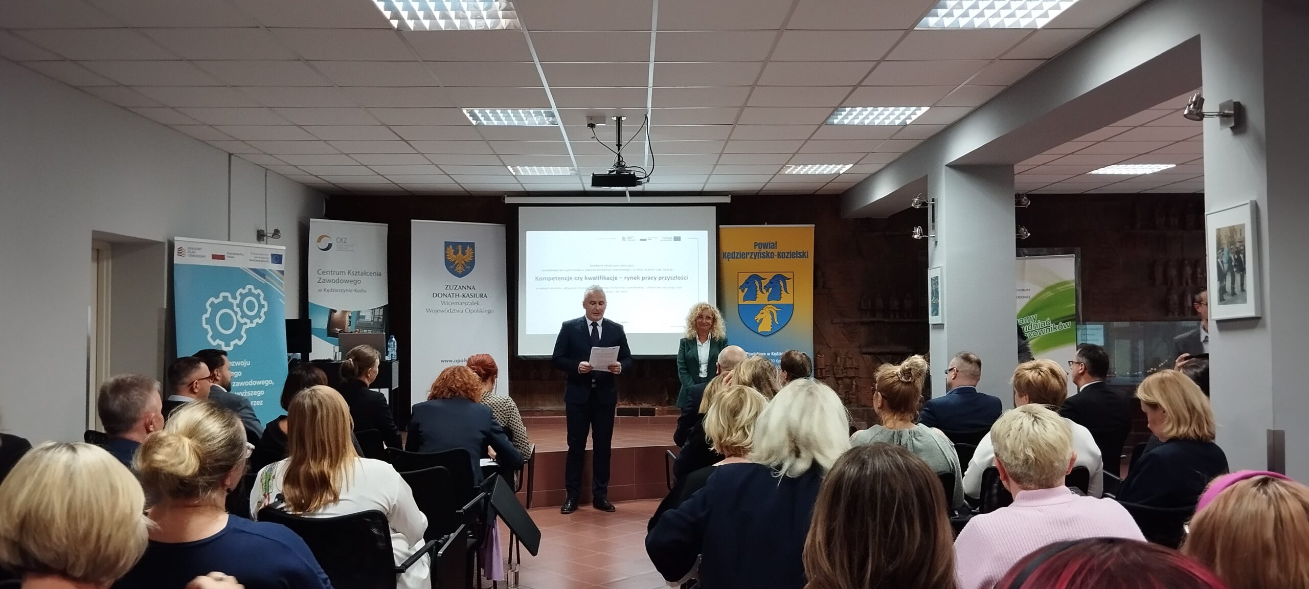 Mężczyzna w garniturze i kobieta stoją z przodu sali, przemawiając do publiczności siedzącej w rzędach. Za nimi znajduje się ekran projektora wyświetlający prezentację, z banerami i plakatami po obu stronach pokoju pod jasnymi światłami sufitowymi.