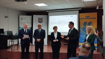 Sześć dorosłych osób stoi na scenie w strojach biznesowych, trzymając dokumenty i rozmawiając. Za nimi znajdują się banery, ekran projektora wyświetlający prezentację i herb. Wygląda to na formalne wydarzenie lub konferencję w profesjonalnym środowisku.