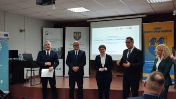 Pięć dorosłych osób stoi na scenie w strojach biznesowych, przemawiając i gestykulując w kierunku publiczności. Za nimi znajdują się ekrany projekcyjne z polskim tekstem, banery i niebieski wyświetlacz z kołami zębatymi. Wydaje się, że jest to oficjalne wydarzenie lub seminarium w Polsce.