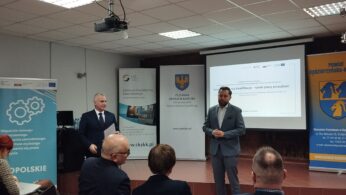 Dwóch mężczyzn w garniturach stoi z przodu sali, prowadząc prezentację dla siedzącej publiczności. Za nimi znajdują się banery, ekran projekcyjny z polskim tekstem i żółto-niebieski emblemat na ścianie. Pomieszczenie ma fluorescencyjne światła i sufit wyłożony kafelkami.