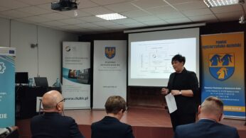 Kobieta w czerni stoi przed uczestnikami konferencji. Za nią na ekranie wyświetlany jest schemat blokowy. Widoczne są banery i znaki z polskim tekstem i logo, a ściany i podłoga są szare i czerwone. Atmosfera jest profesjonalna.