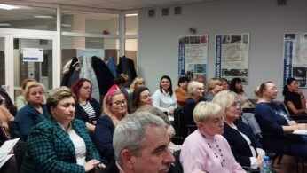 Grupa dorosłych, głównie kobiet, siedzi uważnie w klasie lub sali konferencyjnej z białymi ścianami, plakatami i szklanymi drzwiami. Niektórzy robią notatki, podczas gdy inni słuchają. Atmosfera wydaje się formalna i skupiona.