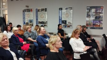 Grupa kobiet i kilku mężczyzn siedzi uważnie w klasie lub sali konferencyjnej, zwróceni twarzami do przodu. Ubrani są w stroje biznesowe lub codzienne. Na białych ścianach za nimi wyświetlane są plakaty i zdjęcia, a niektóre osoby robią notatki.