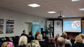 Kobieta stoi z przodu sali konferencyjnej, prowadząc prezentację dla siedzących uczestników. Na ekranie za nią wyświetlany jest pokaz slajdów. Banery i plakaty z tekstem i grafiką są wyświetlane w całym pomieszczeniu, a publiczność uważnie słucha.