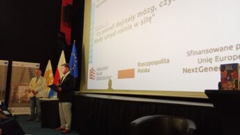 Dwóch mężczyzn stoi na scenie w pobliżu flag i ekranu projekcyjnego pokazującego polski tekst, logo i flagę UE. Jeden mężczyzna trzyma teczkę, drugi stoi z rękami za plecami. Na stole leży książka; na pierwszym planie widoczne są krzesła dla publiczności.