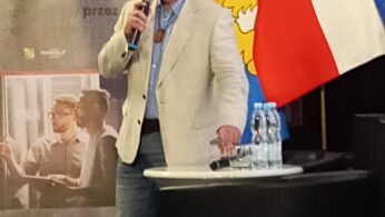 Mężczyzna w jasnej marynarce przemawia do mikrofonu na podium. Za nim znajdują się flagi Polski i UE, żółty emblemat oraz wyświetlacz z tekstem i zdjęciami ludzi. Na podium stoją butelki z wodą. Wydaje się, że jest to formalne wydarzenie lub konferencja.