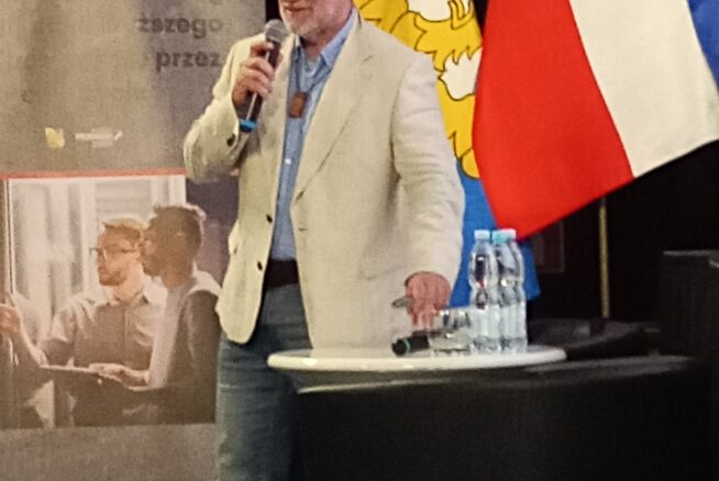 Mężczyzna w jasnej marynarce przemawia do mikrofonu na podium. Za nim znajdują się flagi Polski i UE, żółty emblemat oraz wyświetlacz z tekstem i zdjęciami ludzi. Na podium stoją butelki z wodą. Wydaje się, że jest to formalne wydarzenie lub konferencja.