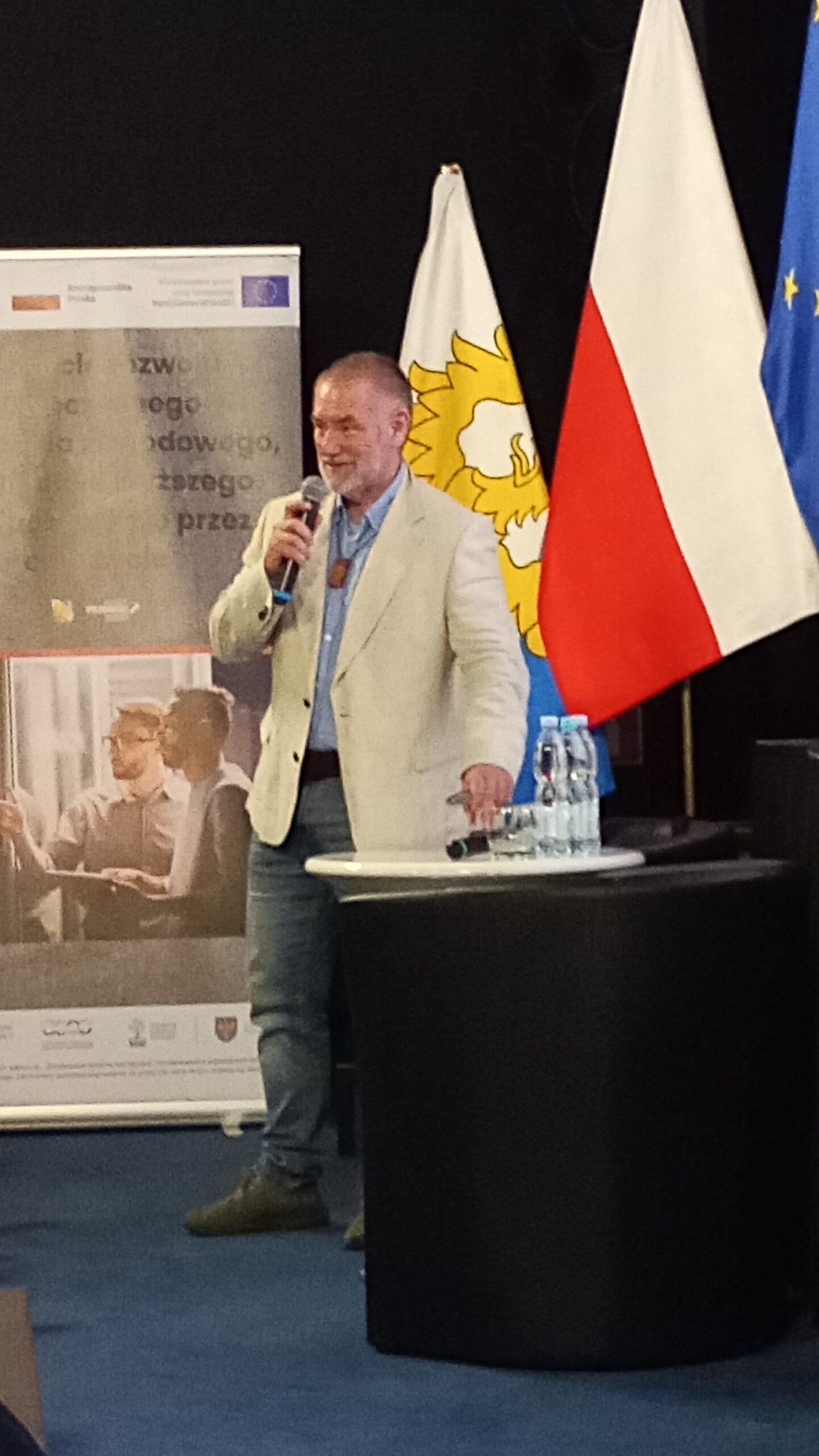 Mężczyzna w jasnej marynarce przemawia do mikrofonu na podium. Za nim znajdują się flagi Polski i UE, żółty emblemat oraz wyświetlacz z tekstem i zdjęciami ludzi. Na podium stoją butelki z wodą. Wydaje się, że jest to formalne wydarzenie lub konferencja.