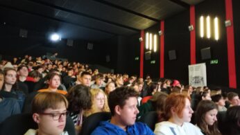 Duża grupa młodych ludzi siedzi w zaciemnionym kinie, twarzą do ekranu. Większość miejsc jest zajęta, a publiczność wydaje się uważna. Ściany mają czerwone pionowe pasy i światła. Po prawej stronie widoczny jest baner z tekstem.