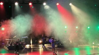 Zespół występuje na scenie wypełnionej kolorowymi światłami i gęstą mgłą. Wielu muzyków gra na instrumentach, podczas gdy wokalista stoi w centrum. Czerwone, zielone i białe reflektory padają w dół, tworząc dramatyczną atmosferę w ciemnym miejscu.