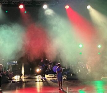 Zespół występuje na scenie wypełnionej kolorowymi światłami i gęstą mgłą. Wielu muzyków gra na instrumentach, podczas gdy wokalista stoi w centrum. Czerwone, zielone i białe reflektory padają w dół, tworząc dramatyczną atmosferę w ciemnym miejscu.