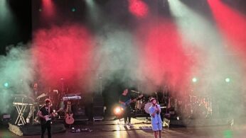 Zespół występuje na scenie pod dramatycznymi czerwono-białymi światłami i ciężką mgłą. Jedna osoba śpiewa do mikrofonu na środku sceny, podczas gdy inni grają na gitarze i klawiszach w tle. Sprzęt muzyczny i instrumenty są widoczne, tworząc żywą scenę koncertową.