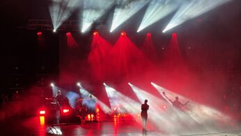Sylwetkowy zespół występuje na scenie wśród dramatycznych czerwonych i białych świateł i dymu, z promieniami świecącymi na zewnątrz. Główny wokalista stoi na środku sceny, podczas gdy muzycy grają na instrumentach. Tłum ludzi ogląda koncert na pierwszym planie.