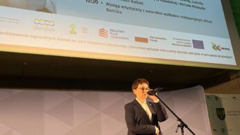 Mężczyzna w garniturze przemawia do mikrofonu na scenie. Za nim znajduje się duży ekran, na którym wyświetlany jest program 