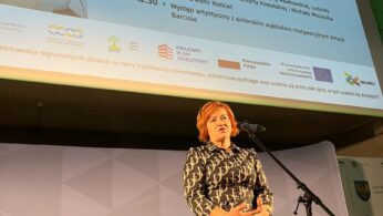 Kobieta we wzorzystej sukience stoi na scenie trzymając mikrofon. Za nią znajduje się duży ekran wyświetlający 