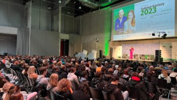 Duża publiczność siedzi w rzędach naprzeciwko sceny, na której na podium przemawia kobieta w różowym garniturze. Za nią jasny ekran wyświetla DoD & Týden kariéry 2023 ze szczegółami wydarzenia i zdjęciami dwóch osób. Pomieszczenie jest nowoczesne z wysokimi sufitami.