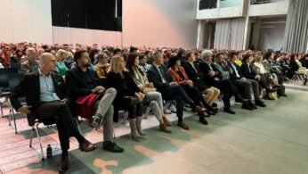 Duża publiczność siedzi uważnie w rzędach krzeseł na imprezie wewnętrznej. Ludzie ubrani są w stroje biznesowe lub eleganckie. Pomieszczenie jest przestronne z wysokimi sufitami, odsłoniętym betonem i gołymi ścianami. Jasne oświetlenie podkreśla skupienie tłumu.