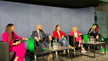 Pięć osób siedzi na scenie w czarnych fotelach, zaangażowanych w dyskusję panelową. Cztery kobiety i jeden mężczyzna stoją twarzą do publiczności. Wszyscy ubrani są w formalne lub biznesowe stroje. Na stołach przed nimi stoi woda butelkowana, a za nimi znajduje się oświetlone tło.