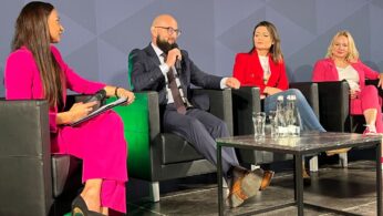 Cztery osoby siedzą na scenie w czarnych fotelach, zaangażowane w dyskusję panelową. Trzy kobiety i jeden mężczyzna, wszyscy w strojach biznesowych, stoją twarzą do publiczności. Kobiety noszą jasnoróżowe stroje, a mężczyzna garnitur. Z przodu stoi szklany stół z butelkami wody i szklankami.