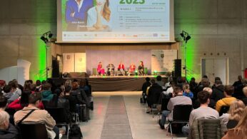 Publiczność ogląda panel czterech mówców na scenie podczas wydarzenia zawodowego w dużej hali. Na ekranie powyżej wyświetlane są szczegóły wydarzenia i zdjęcia dwóch osób. Scena jest oświetlona zielonymi światłami, a publiczność siedzi w rzędach naprzeciwko prelegentów.