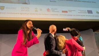 Cztery osoby stoją na scenie podczas wydarzenia, dwie kobiety przytulają się i są ubrane w jasne garnitury, jeden mężczyzna robi sobie z nimi selfie, inna kobieta w różowym garniturze trzyma mikrofon. Za nimi wyświetlany jest duży plakat wydarzenia 