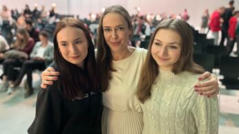 Trzy kobiety stoją blisko siebie, uśmiechając się do kamery. Kobieta w centrum ma na sobie białą sukienkę, a jej ramiona otaczają dwie młodsze kobiety; jedna w czarnej bluzie z kapturem po lewej, a druga w białym swetrze po prawej. Niewyraźna siedząca publiczność jest widoczna w tle.
