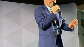 Starszy mężczyzna w niebieskim garniturze, wzorzystym krawacie i okularach przemawia do mikrofonu na scenie, gestykulując lewą ręką. Za nim znajdują się puste czarne krzesła i szare tło z geometrycznym wzorem. Wygląda to na formalne wydarzenie lub konferencję.