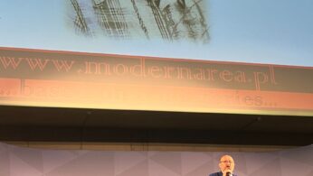 Mężczyzna w garniturze przemawia na scenie, a za nim stoją puste krzesła. Powyżej na dużym ekranie wyświetlany jest czarno-biały portret młodej osoby z prostymi włosami i koszulą w kratę. Na ekranie wyświetlana jest strona internetowa www.modernarea.pl i fraza ...based on true stories....