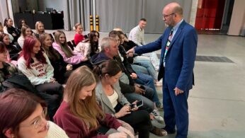 Łysy mężczyzna w niebieskim garniturze trzyma mikrofon w kierunku siwowłosego mężczyzny siedzącego w pierwszym rzędzie widowni. Publiczność składa się głównie z młodych kobiet, niektórych uśmiechniętych i roześmianych, w przestronnym pomieszczeniu z szarymi zasłonami i betonową podłogą.