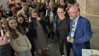 Tłum ludzi, głównie młodych dorosłych, gromadzi się wokół kobiety w okularach i mężczyzny w niebieskim garniturze z kolorowym krawatem, którzy stoją przy stole z książkami. Kilka osób robi zdjęcia, a atmosfera wydaje się żywa i zaangażowana w przestrzeni wewnętrznej.