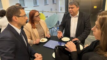 Cztery dorosłe osoby, trzech mężczyzn i jedna kobieta, stoją wokół wysokiego stołu w pomieszczeniu i prowadzą rozmowę. Ubrani są w stroje biznesowe i trzymają małe talerze z ciastkami. Na stole leżą papiery i torebka, a światło z okien rozjaśnia nowoczesne otoczenie.