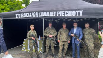 Pięć osób stoi pod czarnym daszkiem z napisem 171 Batalion Lekkiej Piechoty. Cztery osoby mają na sobie kamuflażowe mundury wojskowe i berety, trzymają karabiny. Pośrodku stoi mężczyzna w niebieskim garniturze i okularach. W tle widoczne są drzewa i kostka brukowa.