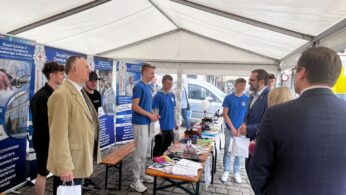 Grupa młodych mężczyzn w niebieskich i czarnych koszulkach stoi za stołem wystawowym pod białym namiotem, prezentując przedmioty kilku dorosłym w garniturach. Za nimi widoczne są plakaty informacyjne z polskim tekstem. Wydarzenie odbywa się na zewnątrz, na brukowanym chodniku.