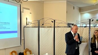 Mężczyzna w garniturze mówi do mikrofonu obok młodej kobiety stojącej przy składanej ściance działowej. Po lewej stronie ekran projekcyjny wyświetla informacje i zdjęcie Justyny Chmielewskiej na profesjonalnym wydarzeniu w jasnym, eleganckim pomieszczeniu.