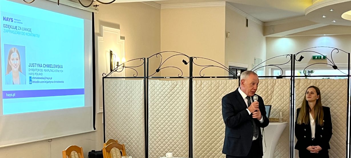 Mężczyzna w garniturze mówi do mikrofonu obok młodej kobiety stojącej przy składanej ściance działowej. Po lewej stronie ekran projekcyjny wyświetla informacje i zdjęcie Justyny Chmielewskiej na profesjonalnym wydarzeniu w jasnym, eleganckim pomieszczeniu.