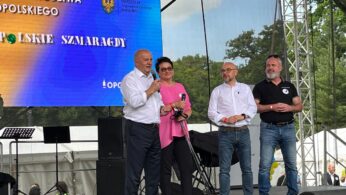 Cztery dorosłe osoby stoją na scenie na świeżym powietrzu przed dużym ekranem z polskim tekstem. Jeden mężczyzna w białej koszuli mówi i gestykuluje. Pozostali, dwóch mężczyzn i kobieta w różowej koszuli, przyglądają się i uśmiechają. W tle widoczne są mikrofony i namioty.
