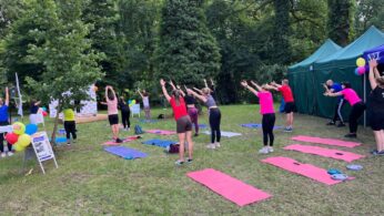 Grupa ludzi wykonuje ćwiczenia fitness na świeżym powietrzu na matach do jogi w trawiastym, zalesionym parku. Większość z nich ma uniesione ramiona i nosi sportowe ubrania. Kolorowe maty i balony zdobią obszar w pobliżu zielonych namiotów. W tle widoczne są znaki i drzewa.