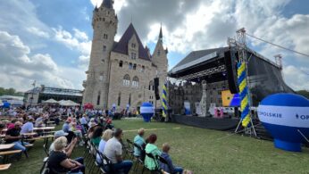 Tłum siedzi na składanych krzesłach naprzeciwko plenerowej sceny udekorowanej niebieskimi i żółtymi balonami. Scena ustawiona jest na trawiastym terenie w pobliżu zamku z wysokimi wieżami. Widocznych jest kilka namiotów i stoisk sprzedawców, a niebo jest częściowo zachmurzone.