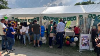 Grupa ludzi stoi przed białym namiotem udekorowanym zielonymi i białymi banerami, balonami i plakatami podczas imprezy plenerowej. Na banerze widnieje napis 