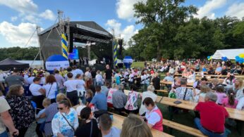 Duża impreza na świeżym powietrzu z wieloma osobami siedzącymi na drewnianych ławkach naprzeciwko sceny. Scena ma czarny baldachim, niebieskie i żółte banery oraz głośniki. W tle widać drzewa i błękitne niebo z chmurami. Niektórzy goście trzymają balony lub noszą odświętne ubrania.