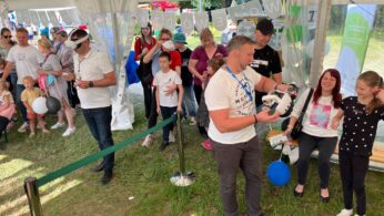 Grupa ludzi, w tym dorośli i dzieci, czeka w kolejce pod białym namiotem. Niektórzy trzymają balony, podczas gdy pracownicy w białych koszulach pomagają i wchodzą w interakcje. Pod stopami widać zieloną trawę, a wewnątrz namiotu wisi baner 