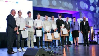 Grupa ośmiu młodych mężczyzn trzymających certyfikaty i torby stoi na scenie między czterema dorosłymi w formalnych strojach, pozując do zdjęcia podczas ceremonii wręczenia nagród. Duży ekran i światła sceniczne są widoczne w nowoczesnym miejscu.