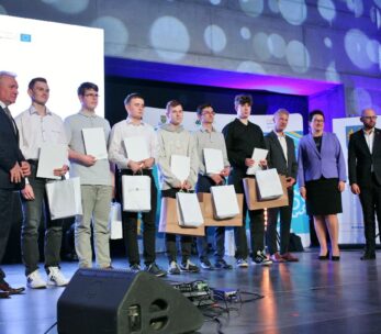 Grupa ośmiu młodych mężczyzn trzymających certyfikaty i torby stoi na scenie między czterema dorosłymi w formalnych strojach, pozując do zdjęcia podczas ceremonii wręczenia nagród. Duży ekran i światła sceniczne są widoczne w nowoczesnym miejscu.