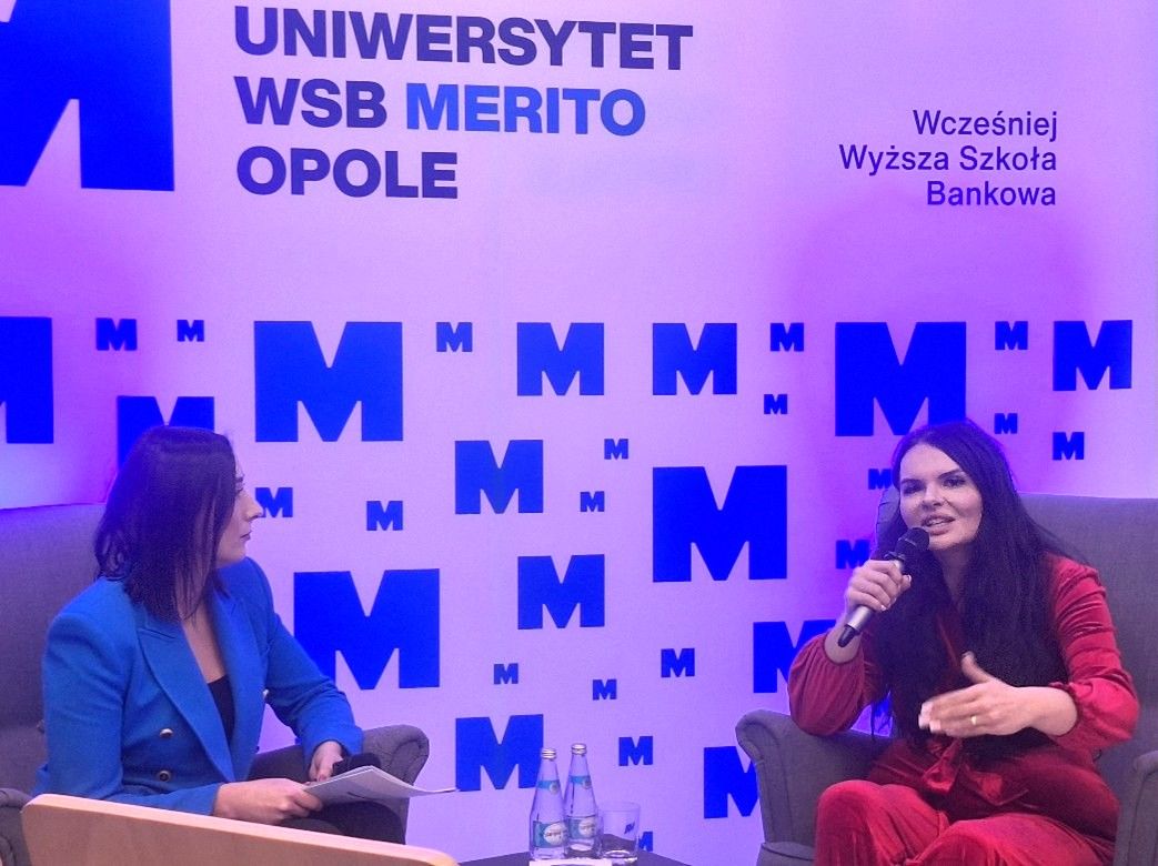 Dwie kobiety siedzą w fotelach na scenie. Kobieta po lewej, w niebieskim garniturze, trzyma notatnik. Kobieta po prawej, w czerwonym kostiumie, mówi do mikrofonu. Za nimi znajduje się ściana z niebieskim logo M i polskim napisem Uniwersytet WSB Merito Opole.