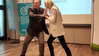Dwoje ludzi, mężczyzna w szarym garniturze i kobieta w białej marynarce, czarnych spodniach i czarnych butach, uśmiechają się i żartobliwie przytulają przed ekranem projekcyjnym w sali konferencyjnej z drewnianą podłogą. Na ekranie wyświetlane są informacje kontaktowe.
