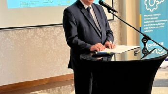 Mężczyzna w ciemnym garniturze stoi na podium z mikrofonem, uśmiechając się podczas przemówienia. Za nim znajduje się ekran projekcyjny wyświetlający polski tekst o konferencji, z logo UE i rządu. Kolejny baner stoi z boku w dobrze oświetlonym pomieszczeniu z wzorzystą podłogą.