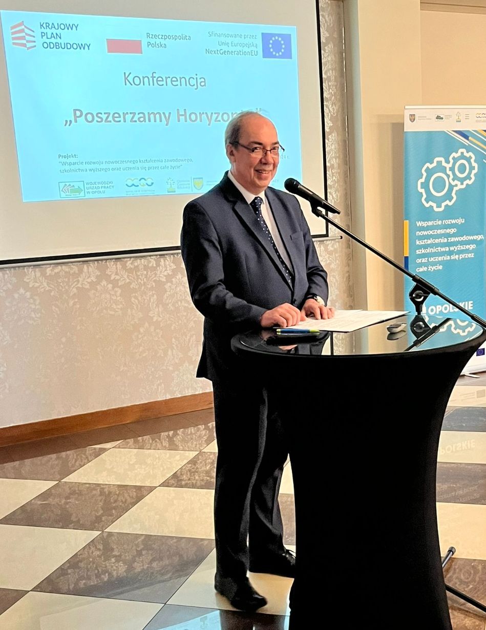 Mężczyzna w ciemnym garniturze stoi na podium z mikrofonem, uśmiechając się podczas przemówienia. Za nim znajduje się ekran projekcyjny wyświetlający polski tekst o konferencji, z logo UE i rządu. Kolejny baner stoi z boku w dobrze oświetlonym pomieszczeniu z wzorzystą podłogą.