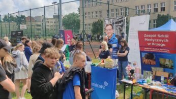 Grupa ludzi stoi na zewnątrz w pobliżu stoisk informacyjnych z plakatami, ulotkami i wystawionymi przedmiotami. Duży napis głosi Zespół Szkół Medycznych w Brzegu. Wydarzenie odbywa się obok boiska sportowego i wielopiętrowego budynku szkoły w pochmurny dzień.