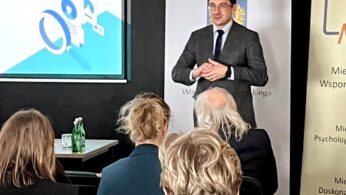 Mężczyzna w garniturze stoi przed ekranem i banerami, przemawiając do publiczności. Cztery osoby siedzą naprzeciw niego. Na ekranie wyświetlana jest niebiesko-biała grafika. Na stole po lewej stronie stoją zielone butelki z wodą. Wygląda to na konferencję lub seminarium.