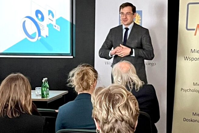 Mężczyzna w garniturze stoi przed ekranem i banerami, przemawiając do publiczności. Cztery osoby siedzą naprzeciw niego. Na ekranie wyświetlana jest niebiesko-biała grafika. Na stole po lewej stronie stoją zielone butelki z wodą. Wygląda to na konferencję lub seminarium.